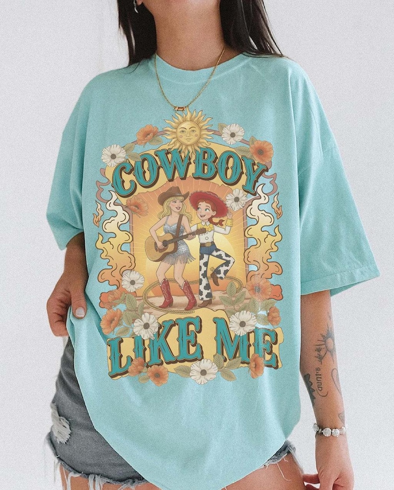 May include: A light blue t-shirt featuring a graphic print with the text "COWBOY LIKE ME" in large blue letters. The design includes a sun, flowers, and two figures, one playing a guitar and the other wearing a cowboy hat, surrounded by flames.