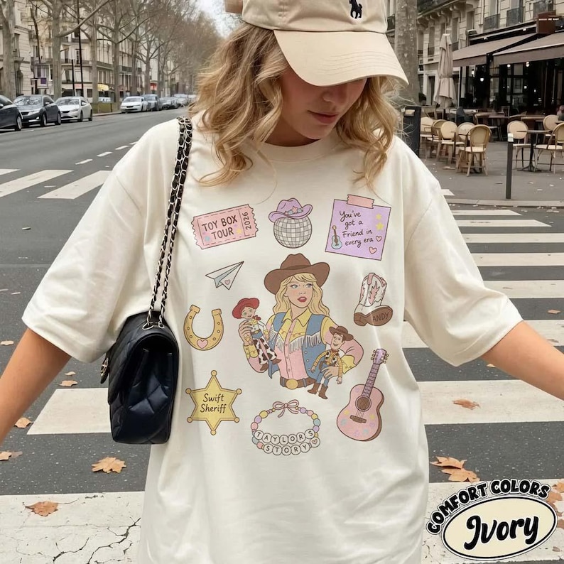 May include: An ivory t-shirt featuring a graphic print of a blonde character in a cowboy hat, a disco ball, a guitar, a horseshoe, a sheriff star labeled Swift Sheriff, a paper airplane, a cowboy boot, and text reading Toy Box Tour 2026.
