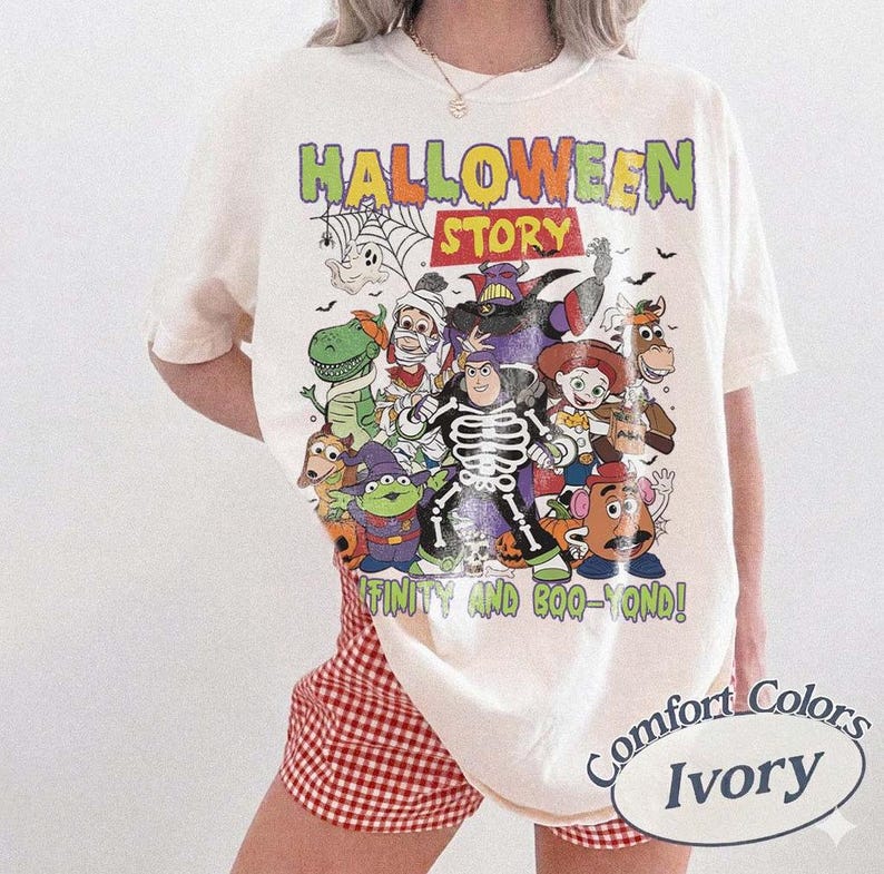 May include: Ivory t-shirt with a colourful graphic featuring cartoon characters and the text "HALLOWEEN STORY" and "INFINITY AND BOO-YOND!". The shirt has a crew neck and short sleeves. The shirt is labelled "Comfort Colors Ivory".