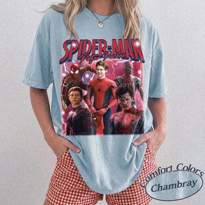 May include: Light blue t-shirt featuring a graphic of Spider-Man and the text "Spider-Man Peter Parker" in red and blue. The shirt has a vintage style and is made of a soft material. The bottom right corner of the shirt has the text "Comfort Colors Chambray".
