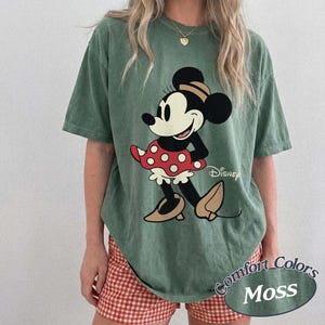 May include: Green t-shirt featuring a Minnie Mouse graphic. The cartoon character is wearing a red polka dot dress and tan shoes. The shirt has the Disney logo and the words "Comfort Colors Moss" printed on it.