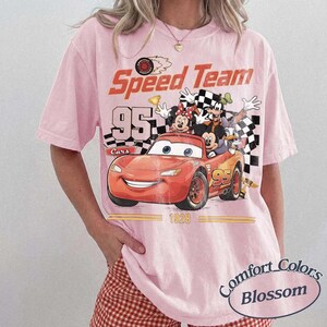 May include: A light pink t-shirt with a graphic featuring a red race car with the number 95, and cartoon characters. The shirt has the words "Speed Team" and "Cars" printed on it. The shirt is a Comfort Colors Blossom.