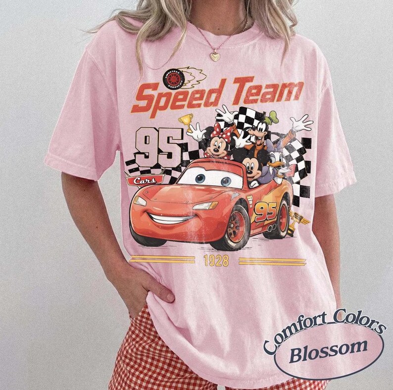 May include: A light pink t-shirt with a graphic featuring a red race car with the number 95, and cartoon characters. The shirt has the words "Speed Team" and "Cars" printed on it. The shirt is a Comfort Colors Blossom.