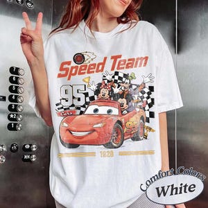 May include: White t-shirt with a graphic featuring the text "Speed Team" and the number "95" with a cartoon car and characters. The shirt also has the text "Cars" and "1928". The shirt is labeled "Comfort Colors White".