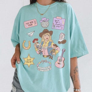 May include: A light blue t-shirt featuring a graphic print of a cowgirl holding two cowboy dolls, surrounded by icons including a disco ball, a guitar, a horseshoe, a sheriff star labeled Swift Sheriff, and text reading Toy Box Tour.