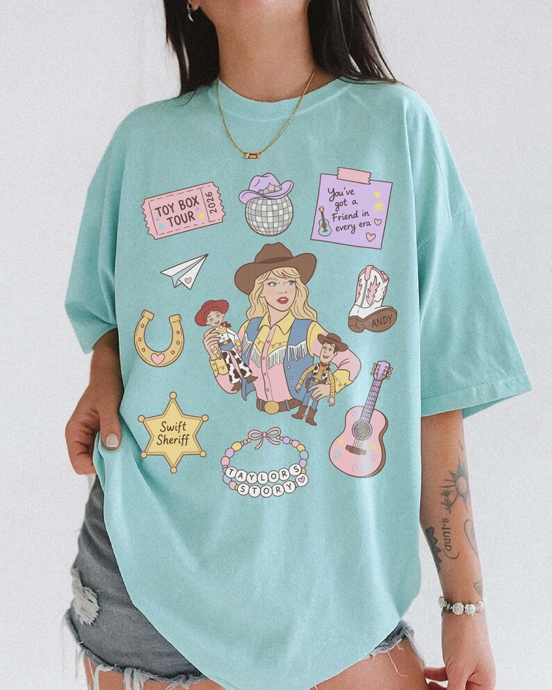 May include: A light blue t-shirt featuring a graphic print of a cowgirl holding two cowboy dolls, surrounded by icons including a disco ball, a guitar, a horseshoe, a sheriff star labeled Swift Sheriff, and text reading Toy Box Tour.