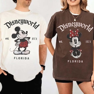 May include: Two short-sleeved t-shirts, one ivory and one espresso, featuring the text "Disneyworld" above a cartoon Mickey Mouse and Minnie Mouse, respectively. The shirts also include "est. 1971" and "Florida" in a vintage style.