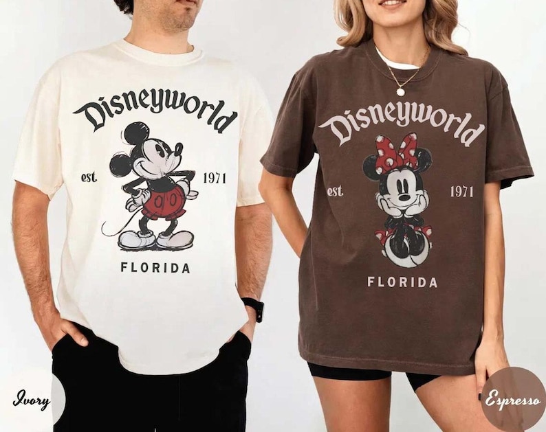 May include: Two short-sleeved t-shirts, one ivory and one espresso, featuring the text "Disneyworld" above a cartoon Mickey Mouse and Minnie Mouse, respectively. The shirts also include "est. 1971" and "Florida" in a vintage style.