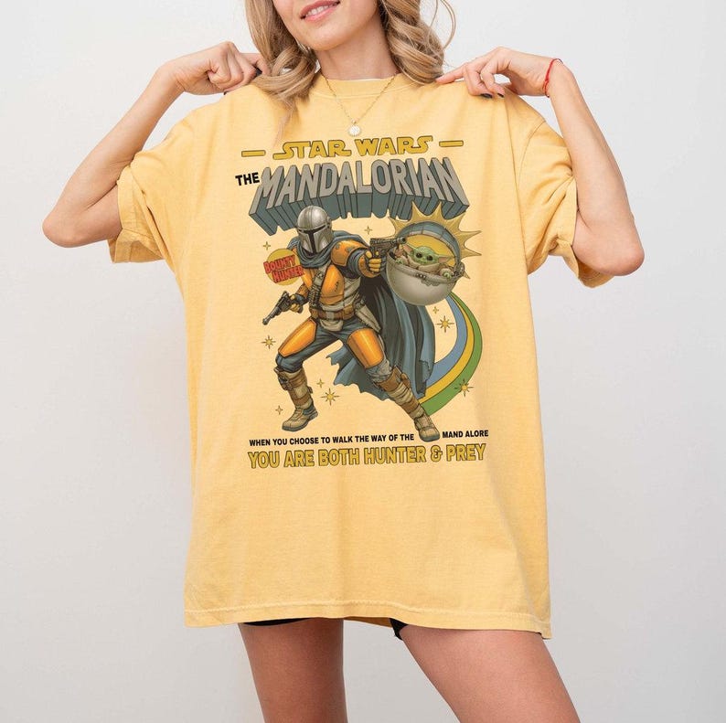 May include: A yellow t-shirt featuring a graphic print of The Mandalorian and Grogu. The text reads Star Wars The Mandalorian, Bounty Hunter, and When you choose to walk the way of the Mandalore you are both hunter and prey with star accents.
