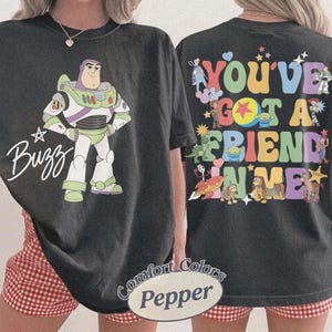 May include: A black t-shirt featuring a graphic of Buzz Lightyear and the text Buzz on the front. The back displays various Toy Story characters and the colorful text You've Got A Friend In Me. The shirt is labeled as Comfort Colors Pepper.