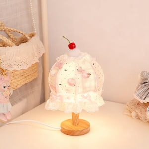 Può includere: Una lampada da tavolo stravagante con una ciliegia in cima. La lampada presenta un paralume in tessuto con un motivo rosa e bianco, adornato con pizzo e fiocchi. Ha una base e uno stelo in legno e un cavo bianco.