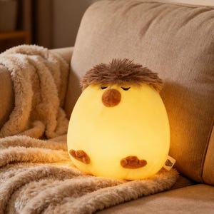 Puede incluir: Una luz de noche amarilla brillante con forma de pájaro con detalles marrones, incluyendo un mechón de pelo, un pico y pies. La luz está sobre una manta beige en un sofá.