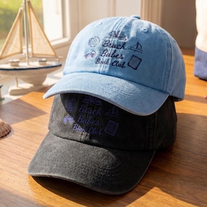 May include: Two baseball caps stacked on top of each other. The top cap is light blue, and the bottom cap is dark gray. Both caps have the embroidered text "Beach Babes Book Club" with seashell, sailboat, and book graphics.