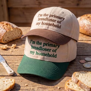May include: Two baseball caps stacked on top of each other. The top cap has a brown brim and the bottom cap has a green brim. Both caps are tan and have the embroidered text, "I'm the primary breadloser of my household."