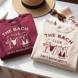 May include: Two crew neck sweatshirts with embroidered text and designs. One is burgundy, the other beige. Both say "THE BACH CLUB" with different names and locations. A straw hat, sunglasses, and a sash are also visible, suggesting a hen party theme.