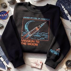 May include: Black crewneck sweatshirt featuring a NASA-themed graphic. The design includes a rocket launching towards a crescent moon, with the text "UNITED STATES OF AMERICA" and "MISSION TO THE MOON." The NASA logo is on the sleeve.