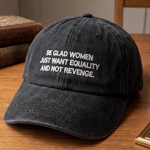 May include: A black baseball cap with white embroidered text that reads "BE GLAD WOMEN JUST WANT EQUALITY AND NOT REVENGE." The cap is made of a soft, worn fabric and has a curved brim. The cap is sitting on a wooden surface.