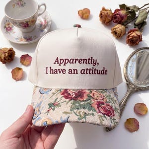 May include: A cream-coloured baseball cap embroidered with the phrase "Apparently, I have an attitude." The brim has a floral pattern in pink, blue, and green. A vintage teacup and saucer are in the background.