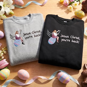 May include: Two sweatshirts, one gray and one black, with the embroidered phrase "Jesus Christ, you're back!" and a cartoon of Jesus on an Easter egg. The sweatshirts are displayed on a wooden surface with Easter decorations.