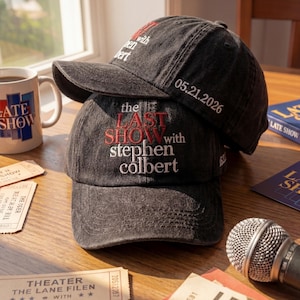 May include: Two black baseball caps stacked on top of each other. The top cap has the text "with en bert" and the date "05.21.2026" embroidered in white. The bottom cap has the text "the LAST SHOW with stephen colbert" embroidered in red and white.