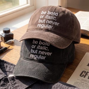 May include: Two baseball caps, one brown and one black, stacked on top of each other. The caps have the embroidered text "be bold or italic, but never regular" in white. The caps are on a wooden surface near an inkwell and a pen.