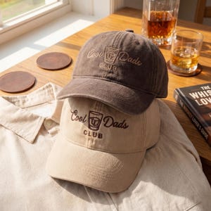 May include: Two baseball caps, one brown and one tan, stacked on top of each other. Both caps have the embroidered text "Cool Dads Club" with a graphic of a cocktail glass. The caps are on a tan button-down shirt.