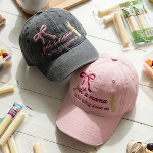 May include: Two baseball caps, one grey and one pink, embroidered with "just a mama in her string cheese era." The caps feature a bow and a string cheese graphic. String cheese and fruit are visible.
