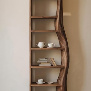 May include: A tall, handcrafted wooden bookshelf with a unique, curved design. The shelves display white teacups, saucers, and a small stack of books. The wood has a rich, warm brown tone, creating a natural and elegant aesthetic.