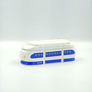 May include: A white and blue ceramic train-shaped container. The container has blue stripes and oval windows. The text "R 67" is printed on the side. The train has a rounded shape and a lid.