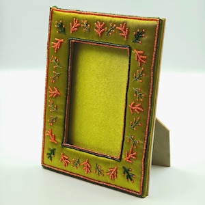 Pode incluir: Uma moldura retangular coberta com tecido de cetim verde-oliva. A moldura apresenta uma borda decorativa de pequenas contas costuradas à mão nas cores coral e verde-escuro, dispostas em um padrão de galhos repetitivo em todos os lados.