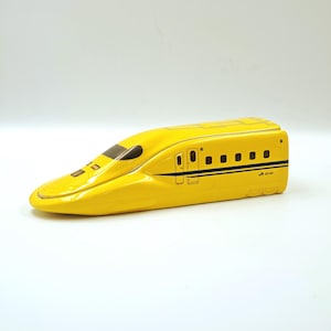 Puede incluir: Modelo de cerámica amarilla de un tren bala japonés, con un diseño aerodinámico, ventanas negras y rayas azules. El tren tiene un frontal detallado y puertas laterales, con el logotipo de JR visible. Sobre un fondo blanco.