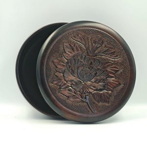 May include: A round, dark brown wooden box with a lid featuring a detailed, carved floral design. The lid displays a prominent flower with layered petals and leaves, set against a textured, stippled background. The box has a smooth, dark interior.
