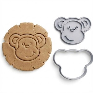 Pode incluir: Um conjunto de cortadores de biscoitos em forma de macaco. O conjunto inclui um biscoito assado com uma cara de macaco impressa e um cortador de plástico cinzento. O biscoito é castanho e o cortador tem a forma de uma cara de macaco.