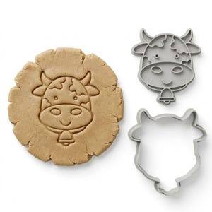 May include: A cow-shaped cookie cutter set. The image shows a pressed cookie with a cow face design and two grey plastic cutters. The cookie is light brown. The cutters are a cow face and a cow outline.