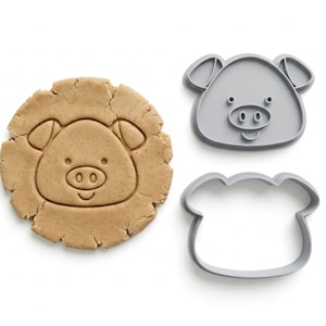 May include: A pig-shaped biscuit and two grey pig-shaped cookie cutters. The biscuit is light brown with a pig face design. The cutters are grey plastic, one in the shape of a pig face and the other in the shape of the pig's head.