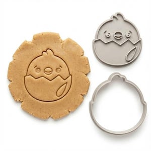 May include: A cookie cutter set featuring a chick hatching from an egg. The set includes a cookie cut-out in dough, a gray plastic cutter in the shape of the chick, and a round cutter. The chick has a smiling face and is light brown.