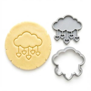 Puede incluir: Una pieza redonda de masa de galleta de color amarillo claro con un diseño de nube en relieve y cinco corazones colgantes. Junto a la masa hay dos cortadores de galletas de plástico gris, uno con el detalle de la nube y otro con la forma.