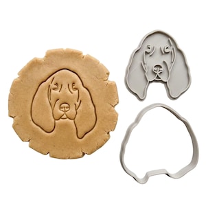 May include: A dog-shaped cookie cutter set with a cookie and a cut-out. The cookie is light brown with an embossed dog face. The cutter is gray plastic, showing the dog's face and the outline for the cookie.