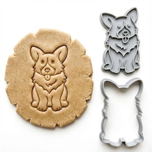 Puede incluir: Un cortador de galletas con forma de corgi y una galleta con forma de corgi se muestran sobre una superficie blanca. El cortador de galletas es de plástico gris y la galleta está hecha de masa marrón. El cortador de galletas tiene dos piezas, una es el contorno y la otra la forma detallada.