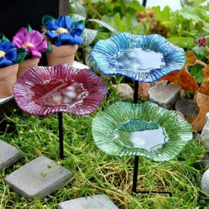 Puede incluir: Tres coloridos bebederos de vidrio en azul, verde y rojo, cada uno con una estaca de metal negro. Los platos están colocados en un jardín con follaje verde y pequeñas piedras grises. Hay macetas decorativas en el fondo.