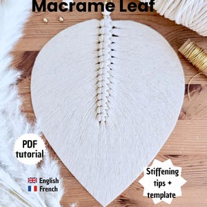 Op de afbeelding: Een handgemaakte macramé blad wanddecoratie in gebroken wit. Het bladontwerp heeft een centrale, getextureerde ruggengraat en een glad, waaiervormig patroon. De tekst "Macrame Leaf" staat bovenaan. Extra tekst: "PDF tutorial", "English French", en "Stiffening tips + template".