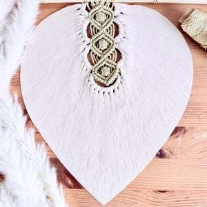 Peut inclure: Décoration murale blanche en forme de cœur avec une surface texturée et un motif macramé au centre. Le macramé est de couleur beige clair. La décoration est suspendue à une cheville en bois avec une corde naturelle.