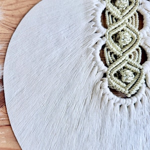 Pode incluir: Macram&eacute; de parede creme com um design circular. A pe&ccedil;a apresenta uma superf&iacute;cie texturizada e com franjas e detalhes intrincados em n&oacute;s em um tom creme mais claro. Uma vareta de madeira sustenta a parte superior.