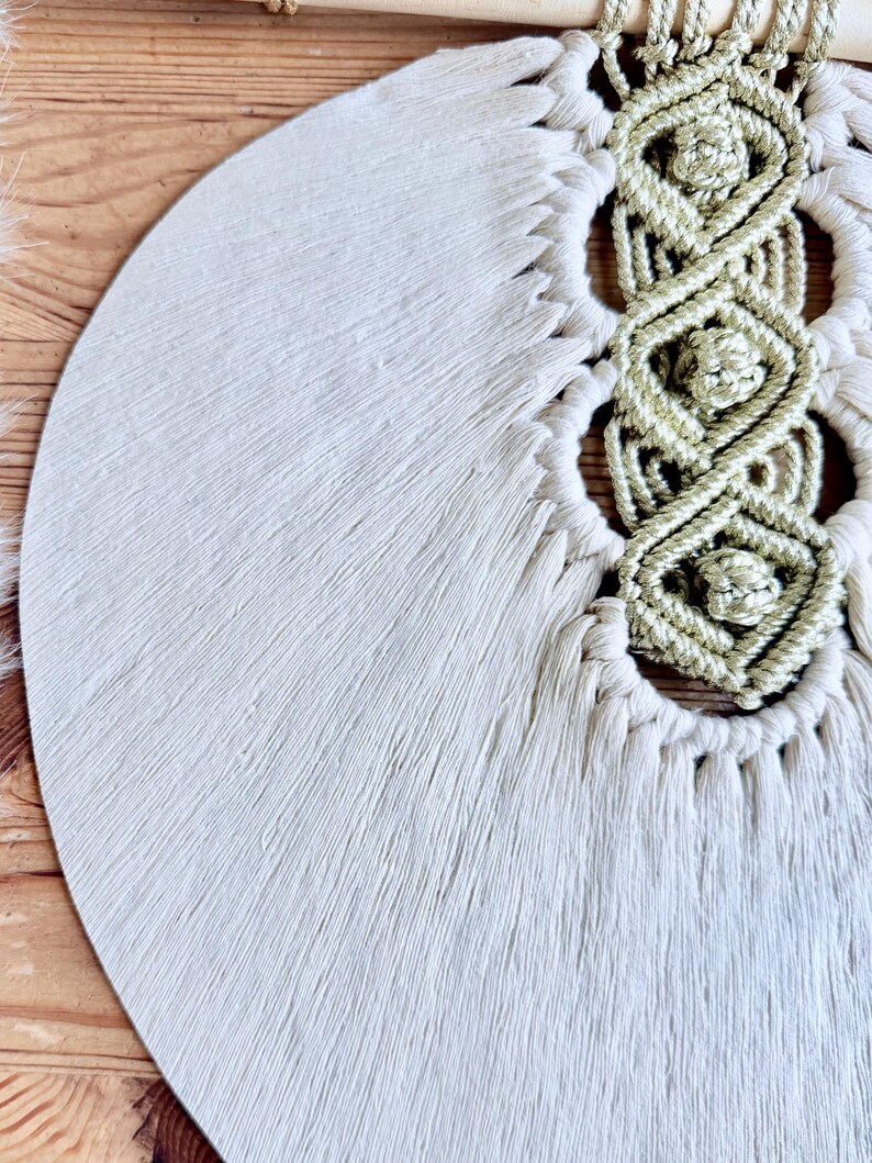 Pode incluir: Macram&eacute; de parede creme com um design circular. A pe&ccedil;a apresenta uma superf&iacute;cie texturizada e com franjas e detalhes intrincados em n&oacute;s em um tom creme mais claro. Uma vareta de madeira sustenta a parte superior.