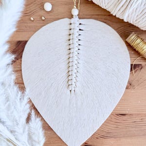 以下が含まれることがあります： クリーム色のマクラメの葉の形をした壁掛け。中央の背骨はしっかりと結ばれ、葉の端が広がっています。吊り下げ用の金色のコードとビーズが上部にあります。背景には木製の表面と白い羽が含まれています。