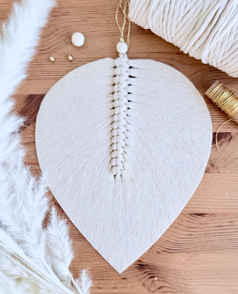 以下が含まれることがあります： クリーム色のマクラメの葉の形をした壁掛け。中央の背骨はしっかりと結ばれ、葉の端が広がっています。吊り下げ用の金色のコードとビーズが上部にあります。背景には木製の表面と白い羽が含まれています。
