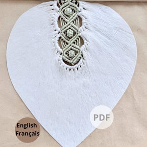 Pode incluir: Uma decora&ccedil;&atilde;o de parede em forma de cora&ccedil;&atilde;o cinza claro com um design de macram&eacute; no centro. O macram&eacute; &eacute; de cor verde claro e &eacute; preso a um pino de madeira. A decora&ccedil;&atilde;o de parede tem uma apar&ecirc;ncia macia e texturizada. As palavras "English Fran&ccedil;ais" s&atilde;o impressas no canto inferior esquerdo.