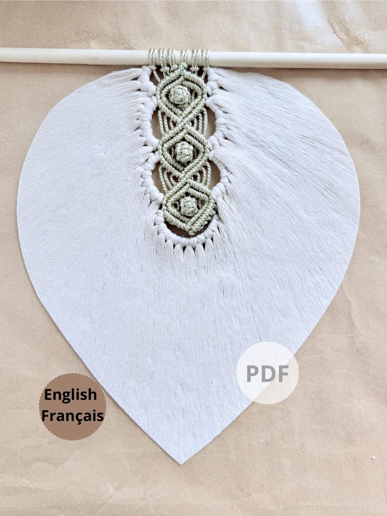 Pode incluir: Uma decora&ccedil;&atilde;o de parede em forma de cora&ccedil;&atilde;o cinza claro com um design de macram&eacute; no centro. O macram&eacute; &eacute; de cor verde claro e &eacute; preso a um pino de madeira. A decora&ccedil;&atilde;o de parede tem uma apar&ecirc;ncia macia e texturizada. As palavras "English Fran&ccedil;ais" s&atilde;o impressas no canto inferior esquerdo.