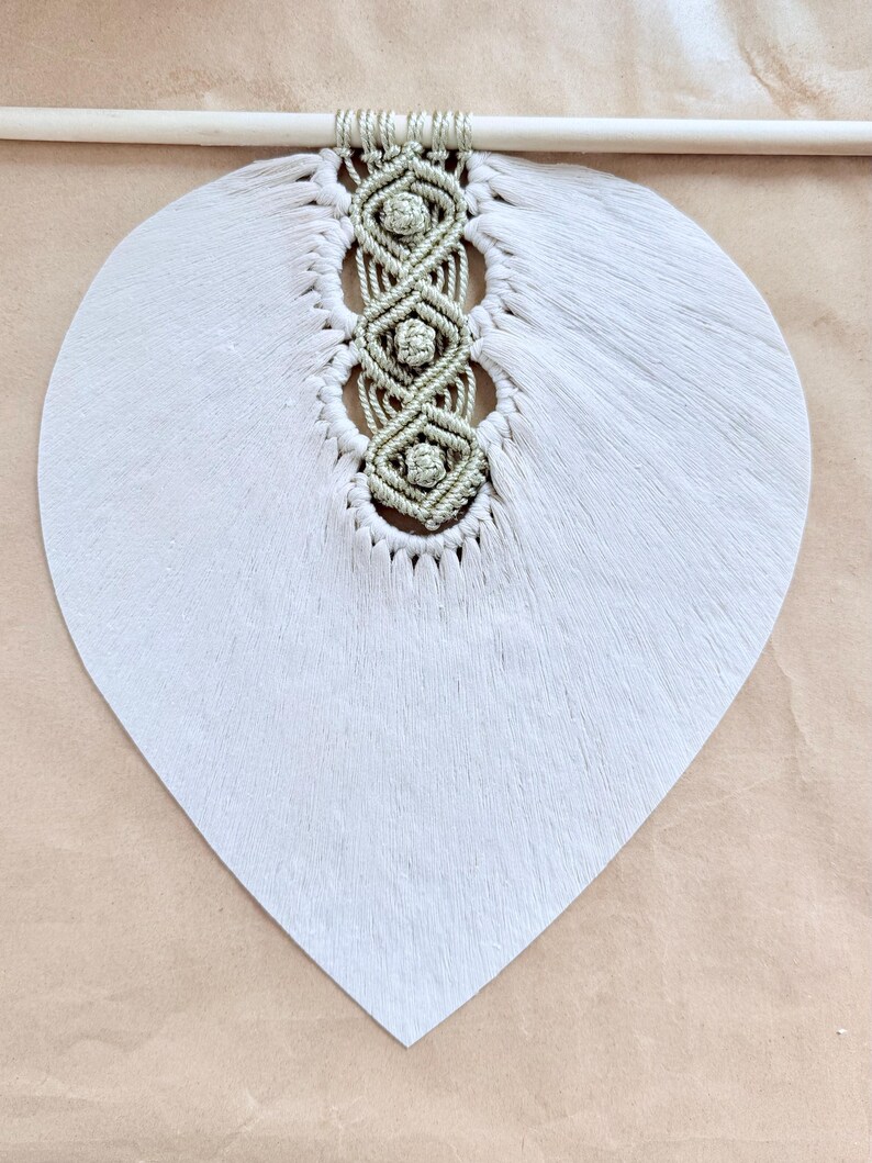 Pode incluir: Um pendurador de parede artesanal com um design grande em forma de folha cinza claro. O centro tem um padr&atilde;o de macram&eacute; em uma cor verde suave, com um pino de madeira na parte superior para pendurar. A textura da folha &eacute; vis&iacute;vel.