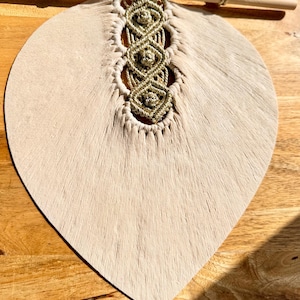 Pode incluir: Um pendurador de parede bege em forma de folha com um design de macram&eacute; dourado. O macram&eacute; &eacute; preso a um pino de madeira. O pendurador de parede &eacute; feito de um material macio e texturizado, suspenso por um fino cord&atilde;o bege.